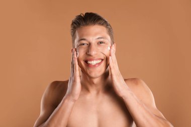 Yakışıklı genç adam açık kahverengi arka planda yüzüne krem sürüyor.