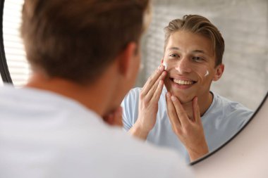 Yakışıklı genç adam evde aynanın yanında yüzüne krem sürüyor.