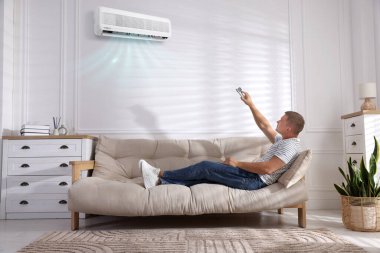 Evdeki kanepede uzaktan kumandayla klima kullanan bir adam.