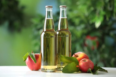 Cam şişelerde lezzetli elma şarabı ve açık havada beyaz ahşap masada elmalar.