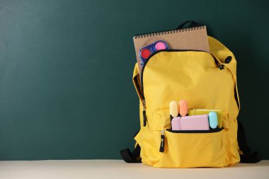 Tahtanın yanındaki masada farklı kırtasiye malzemesi olan sarı sırt çantası. Metin için boşluk