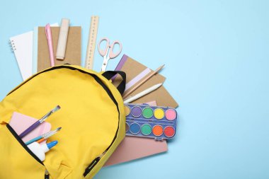 Açık mavi zemin üzerinde farklı kırtasiye malzemesi olan sarı sırt çantası, üst manzara. Metin için boşluk