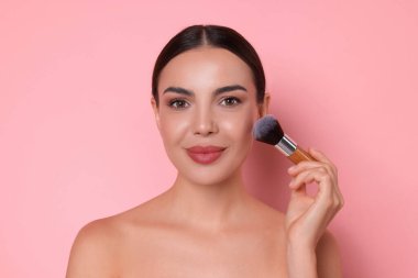 Güzel bir kadın pembe arka planda fırçayla makyaj yapıyor.