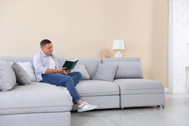 Genç adam evdeki kanepede kitap okuyor.