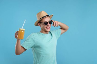 Güneş gözlüklü, şapkalı, açık mavi arka planda portakal suyu kavanozu olan mutlu bir adam. Tazeleyici içecek.