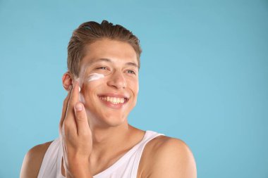 Yakışıklı genç adam yüzüne krem sürüyor açık mavi arka planda, mesaj için yer var.