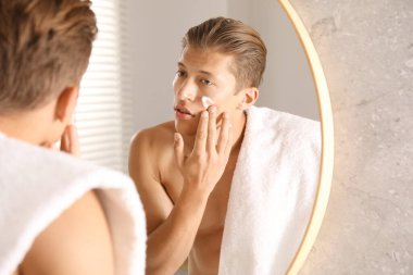 Yakışıklı genç adam banyoda aynanın yanında yüzüne krem sürüyor.