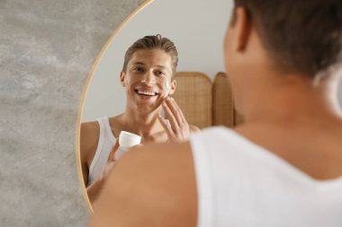 Yakışıklı genç adam banyoda aynanın yanında yüzüne krem sürüyor.