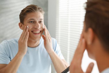 Yakışıklı genç adam evde aynanın yanında yüzüne krem sürüyor.