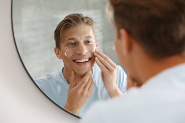 Yakışıklı genç adam evde aynanın yanında yüzüne krem sürüyor.