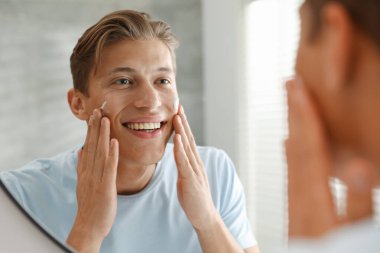 Yakışıklı genç adam evde aynanın yanında yüzüne krem sürüyor.