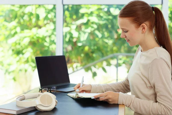 Not defterine yazan ve içerideki ahşap masada dizüstü bilgisayar kullanan genç bir kız. Ev işyeri