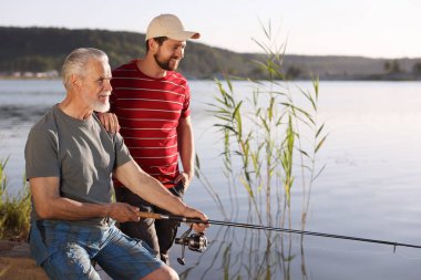 Yaşlı babası ve mutlu oğlu birlikte nehir kenarında balık tutuyorlar.