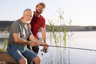Yaşlı babası ve mutlu oğlu birlikte nehir kenarında balık tutuyorlar.