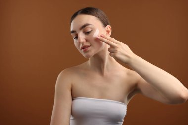 Güzel bir kadın kahverengi arka planda yüzüne krem sürüyor.
