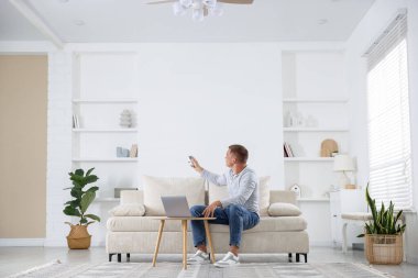 Evdeki kanepede uzaktan kumandayla klima kullanan bir adam.