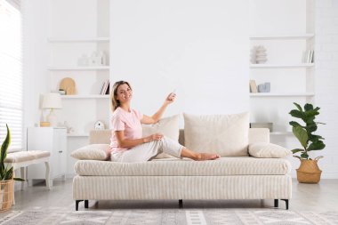 Evdeki koltukta uzaktan kumandayla klima kullanan bir kadın.