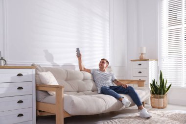 Evdeki kanepede uzaktan kumandayla klima kullanan bir adam.