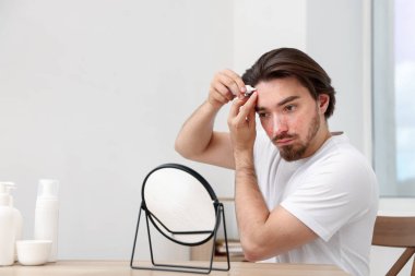 Akne sorunu olan genç bir adam evdeki masada aynanın yanında yüzüne krem sürüyor.