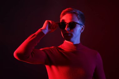 Güneş gözlüğü takan yakışıklı adam karanlık arka plana karşı neon ışıklarla bakıyor.