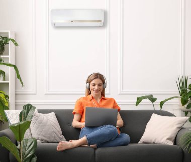 Kulaklıklı bir kadın evdeki kanepede dizüstü bilgisayar kullanıyor. Klima odada rahat bir sıcaklık sağlıyor.