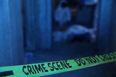 Adli tıp uzmanları terk edilmiş binada cesetle birlikte olay yerinde çalışıyorlar. Sarı polis bandına odaklanıyorlar.