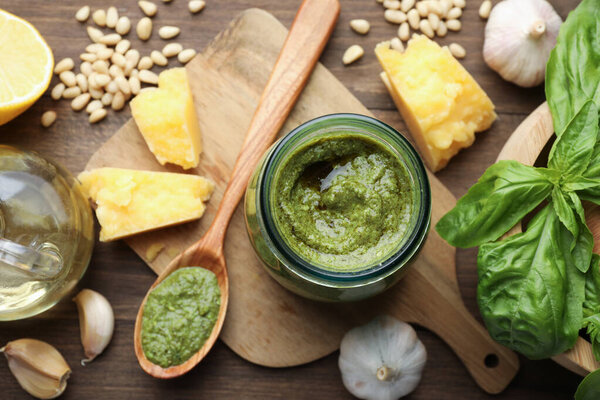 Fresh pesto sauce in jar and ingredients on wooden table, flat lay