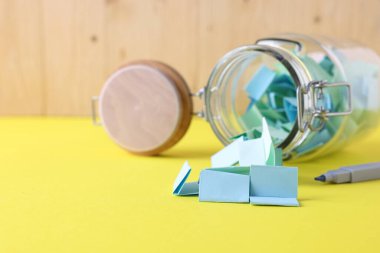 Cam kavanozdaki renkli kağıt notlar ve sarı masadaki keçeli kalem, yakın plan. Metin için boşluk