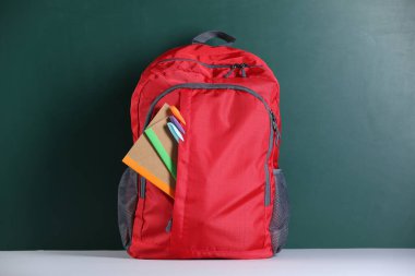 Tahtanın yanındaki masada farklı kırtasiye malzemesi olan kırmızı sırt çantası.