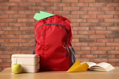 Sırt çantası, kağıt uçaklar, kitap, beslenme çantası ve elma kırmızı tuğla duvara karşı ahşap masada.