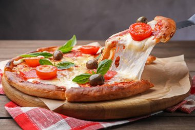 Taking piece of delicious pizza with cheese, salami, tomatoes and olives at wooden table, closeup
