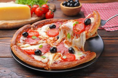 Taking piece of delicious pizza with cheese, salami, tomatoes and olives at wooden table, closeup