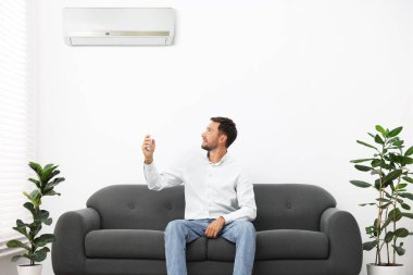 Evdeki kanepede uzaktan kumandayla klima kullanan bir adam.