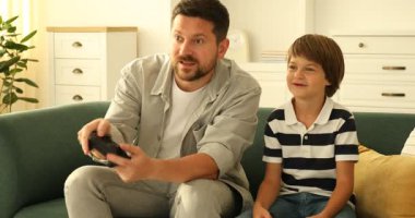 Evde kanepede video oyunu oynayan kontrolörlü baba ve oğul.