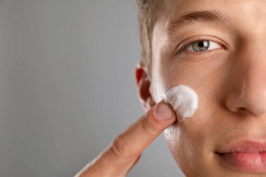Gri arka planda yüzüne krem süren yakışıklı genç adam, yakın plan. Metin için boşluk