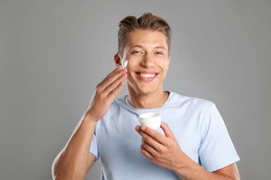 Yakışıklı genç adam gri arka planda yüzüne krem sürüyor.