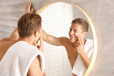 Yakışıklı genç adam banyoda aynanın yanında yüzüne krem sürüyor.