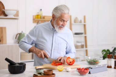 Mutfaktaki beyaz mermer masada yemek pişirirken tablet kullanan yaşlı adam.