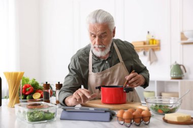 Mutfaktaki beyaz mermer masada yemek pişirirken tablet kullanan yaşlı adam.