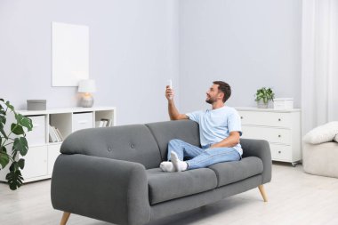 Evdeki kanepede uzaktan kumandayla klima kullanan bir adam.