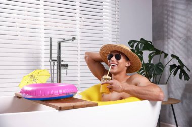 Güneş gözlüklü ve hasır şapkalı mutlu adam evdeki küvette şişme yüzüklü mason kavanozundan meyve suyu içiyor. Denizde tatilin hayalini kuruyorum.