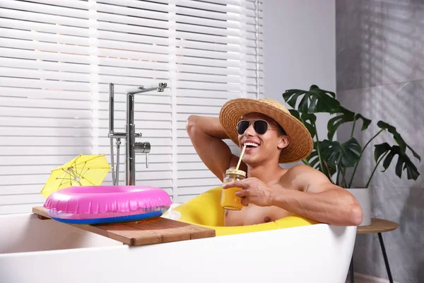 Güneş gözlüklü ve hasır şapkalı mutlu adam evdeki küvette şişme yüzüklü mason kavanozundan meyve suyu içiyor. Denizde tatilin hayalini kuruyorum.