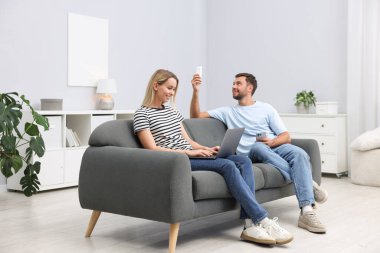 Dizüstü bilgisayarı ve akıllı telefonu olan çift evdeki kanepede klimayı kontrol etmek için uzaktan kumanda kullanıyor.