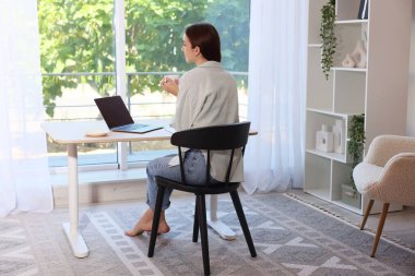 Kahveli bir kadın, ahşap masa arkasında dizüstü bilgisayar kullanıyor. Ev işyeri