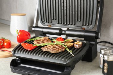 Açık gri masada biftek, biberiye, sebze ve baharatla elektrikli ızgara, yakın plan.