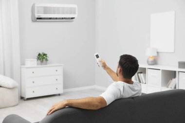 Evdeki kanepede uzaktan kumandayla klima kullanan bir adam.