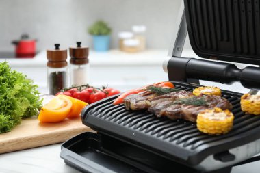 Mutfaktaki beyaz mermer masada lezzetli et, sebze ve baharatlarla elektrikli ızgara, yakın plan.