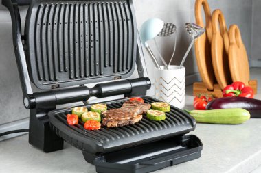 Open electric grill with beef steak, vegetables and cookware on light grey table, closeup
