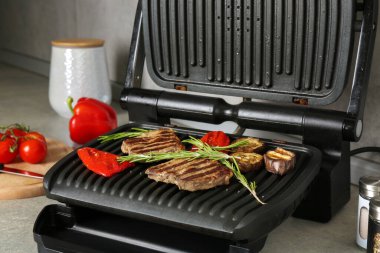 Open electric grill with beef steaks, rosemary, bell pepper and eggplant on light grey table, closeup