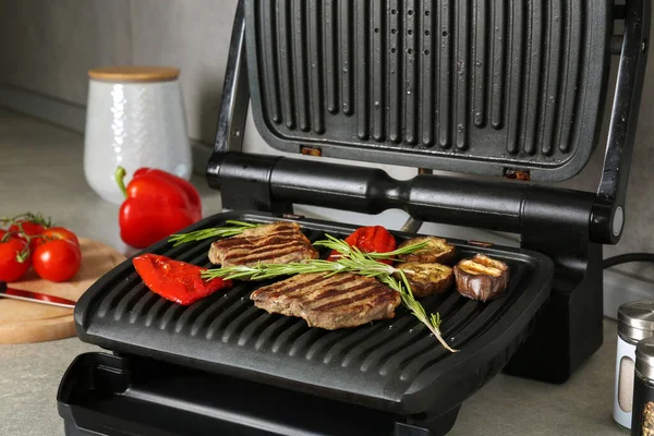Open electric grill with beef steaks, rosemary, bell pepper and eggplant on light grey table, closeup